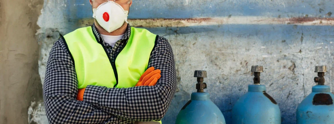 Un trabajador de la construcción masculino de pie con los brazos cruzados, vestido con equipo de protección individual completo, que incluye casco amarillo, gafas de seguridad, mascarilla respiratoria con válvula roja y chaleco de alta visibilidad sobre una camisa a cuadros, delante de una pared azul y tres bombonas de gas azules industriales.