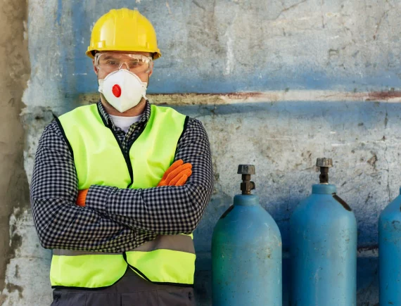 Un trabajador de la construcción masculino de pie con los brazos cruzados, vestido con equipo de protección individual completo, que incluye casco amarillo, gafas de seguridad, mascarilla respiratoria con válvula roja y chaleco de alta visibilidad sobre una camisa a cuadros, delante de una pared azul y tres bombonas de gas azules industriales.